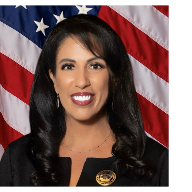 Professional portrait of a woman, wearing dark colored business attire, against a blue backdrop and American flag.