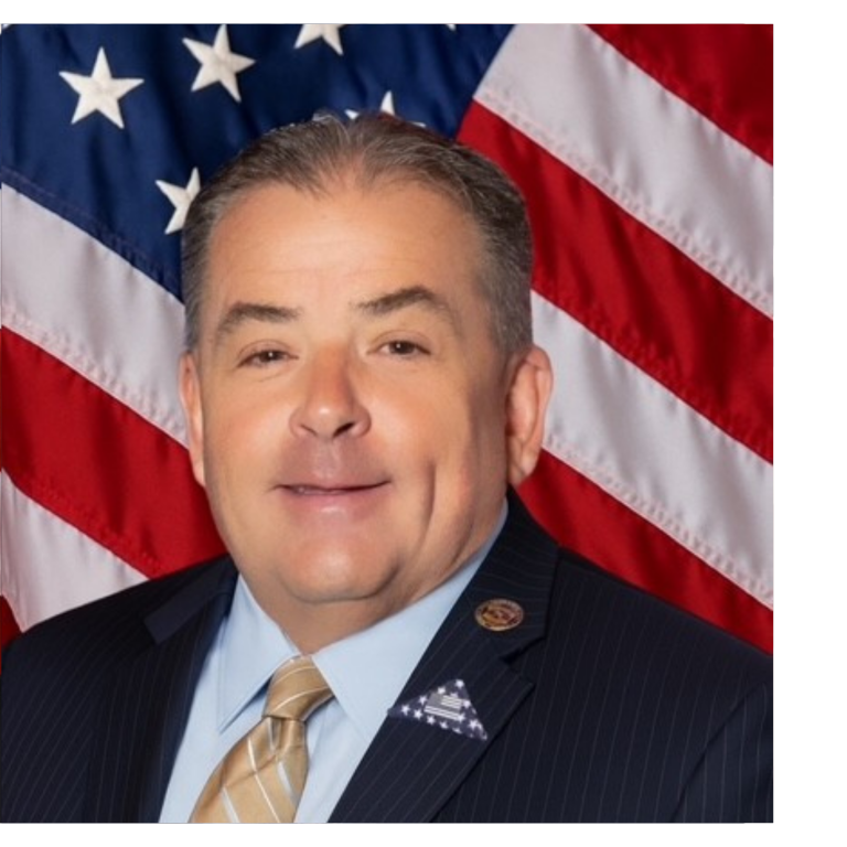 Professional portrait of a man, wearing dark colored business attire, against a blue backdrop and American flag.