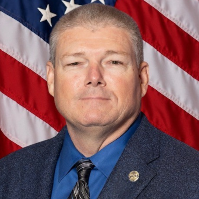 Professional portrait of a man, wearing dark colored business attire, against a blue backdrop and American flag.