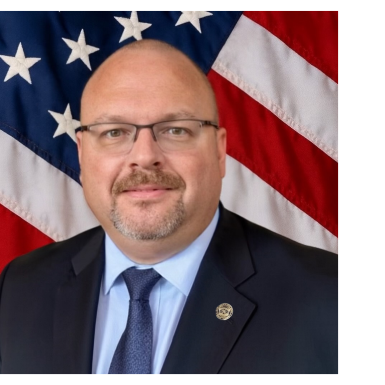 District 3 LEL Brian Muhlbach Professional portrait of a man, wearing dark colored business attire, against a blue backdrop and American flag.
