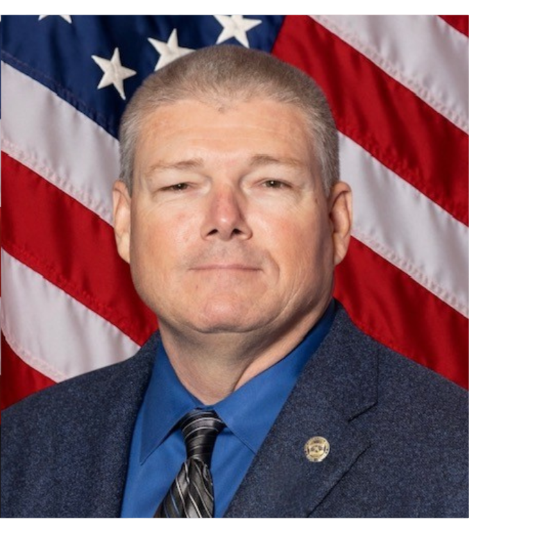 Professional portrait of a man, wearing dark colored business attire, against a blue backdrop and American flag.