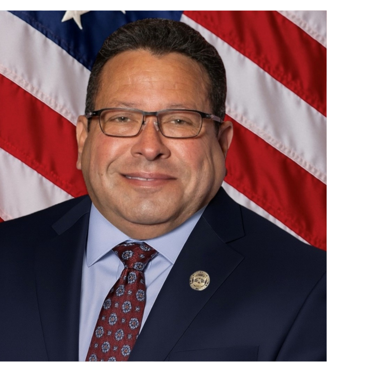 Professional portrait of a man, wearing dark colored business attire, against a blue backdrop and American flag.