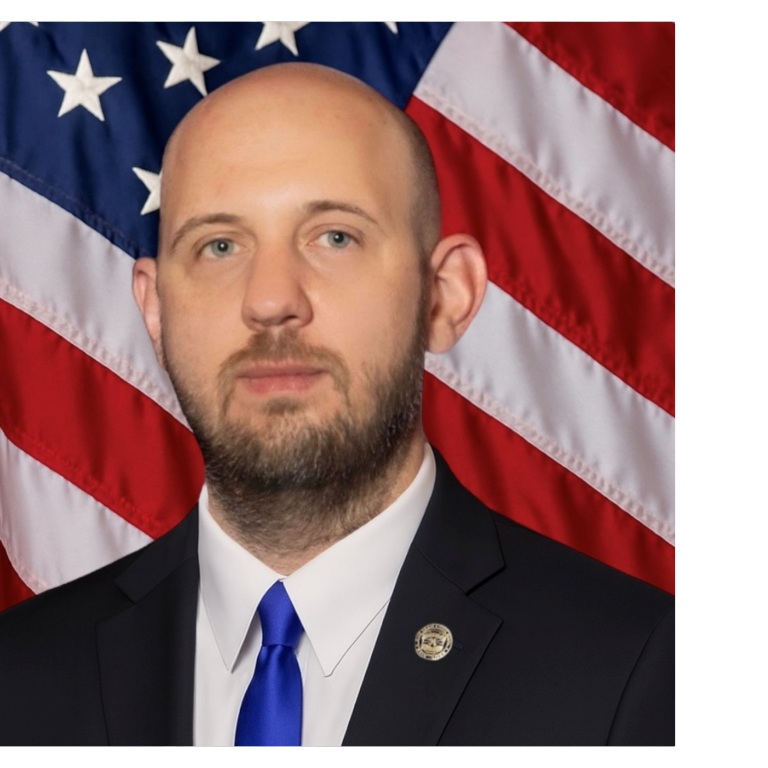 Professional portrait of a man, wearing dark colored business attire, against a blue backdrop and American flag.