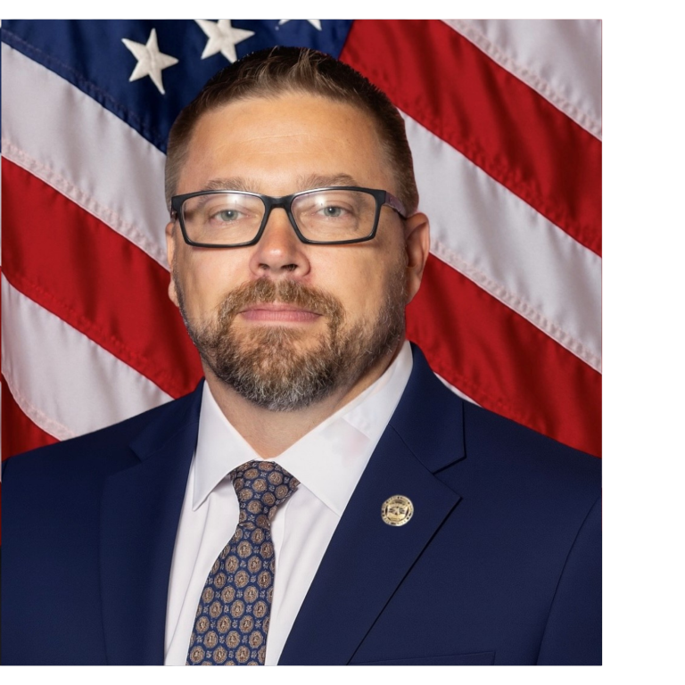 Professional portrait of a man, wearing dark colored business attire, against a blue backdrop and American flag.